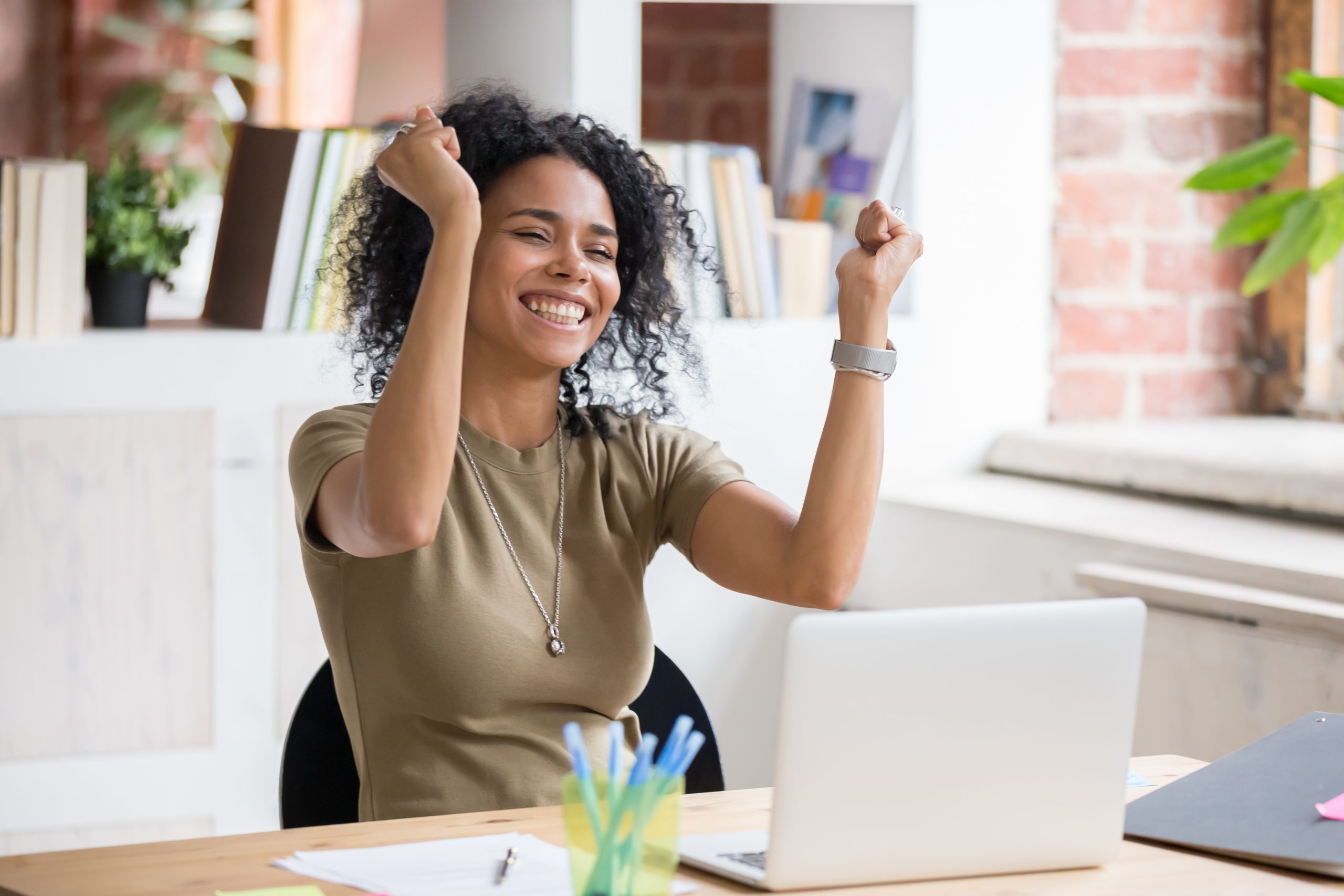 A woman at a computer smiling and raising her hands in celebration