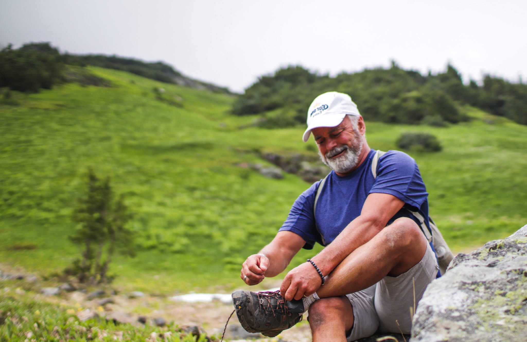 An older man on a hike in the hills, sits to tie his boot lace