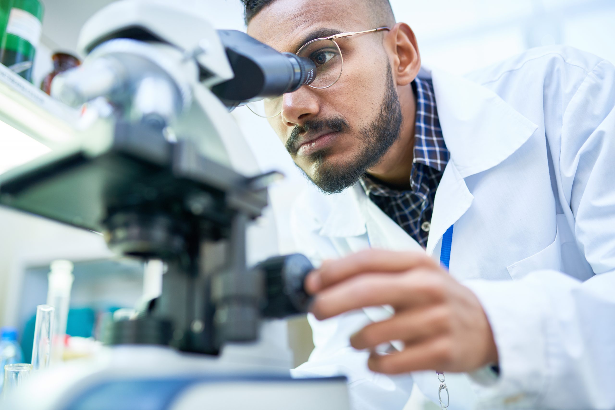 A bearded man with glasses gazes into a microscope in a lab
