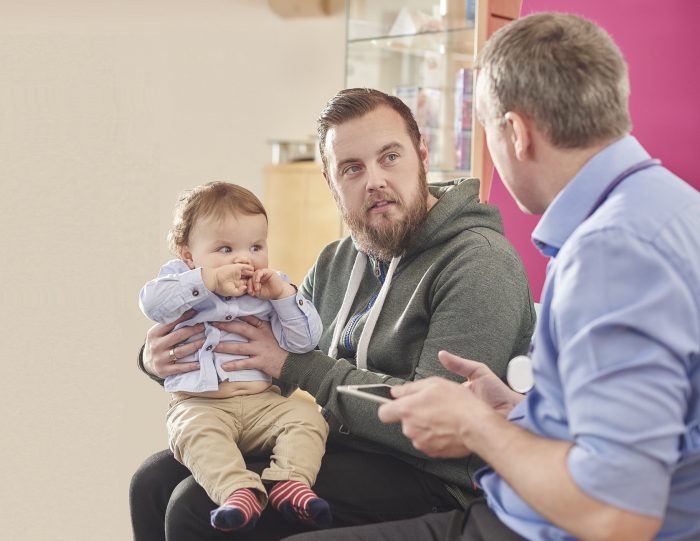A father with beard and a baby seated on his knee, speaks to a male doctor