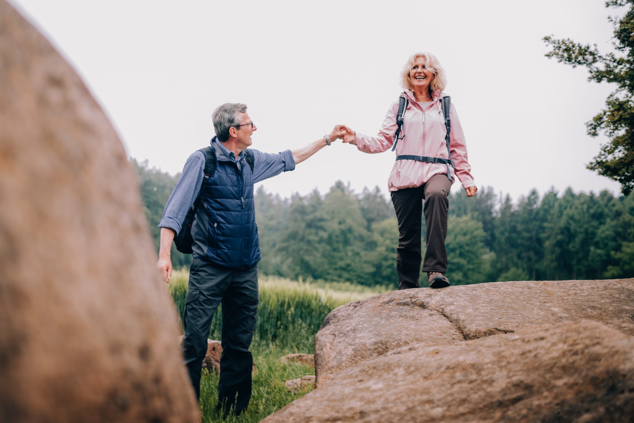 A middle aged man holds his wife's hand as they traverse rocks on a hike